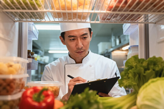 A focused Asian male chef in uniform checks and records ingredients inside a commercial restaurant refrigerator in a professional kitchen.
