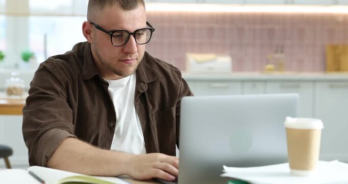 Workaholic. Overwhelmed man wearing glasses working with laptop at desk in kitchen, camera moving
