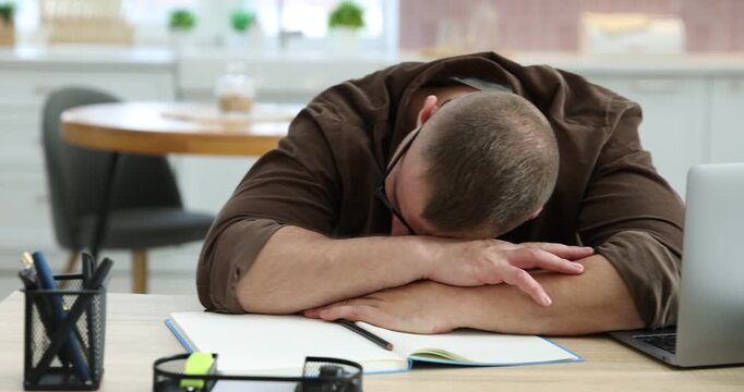 Workaholic. Overwhelmed man in glasses sleeping at wooden desk with laptop and notebook in kitchen, camera moving left