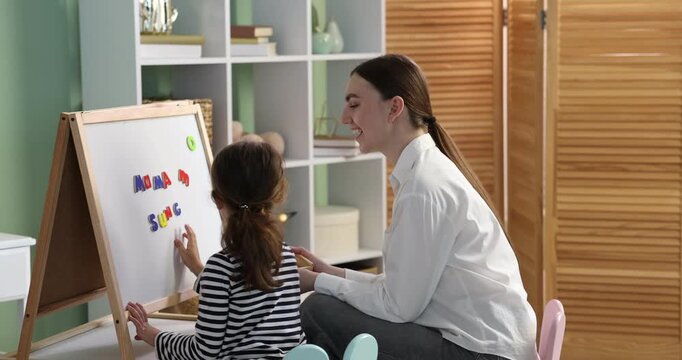 Speech therapist teaching girl alphabet with magnetic letters near whiteboard indoors