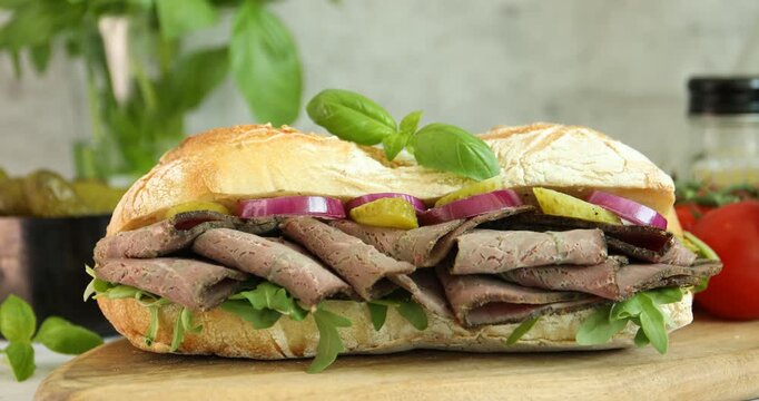 Woman adding basil to delicious sandwich with pastrami (cured beef meat), pickles and onion at table, closeup
