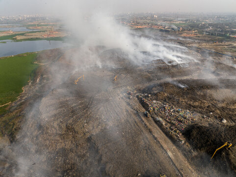 Aminbazar, Bangladesh - 21 February 2026: Aerial view of the Aminbazar landfill with thick smoke rising from waste piles, yellow excavators at work, and a hazy city skyline.