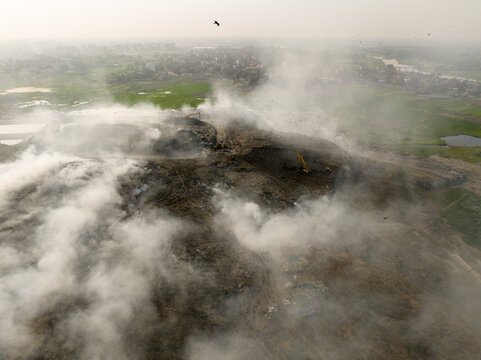 Aminbazar, Bangladesh - 21 February 2026: Aerial view of Aminbazar landfill with thick smoke rising from waste piles, excavators operating, and birds circling over the site.