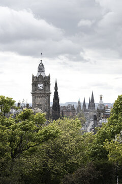 View of the Balmoral Hotel clock tower and Scott Monument rising above lush green trees under a cloudy sky in Edinburgh, Scotland, United Kingdom.