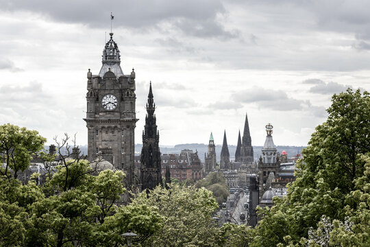 View of the historic skyline featuring the Balmoral Hotel clock tower and Scott Monument framed by lush green trees under a cloudy sky in Edinburgh, Scotland, United Kingdom.