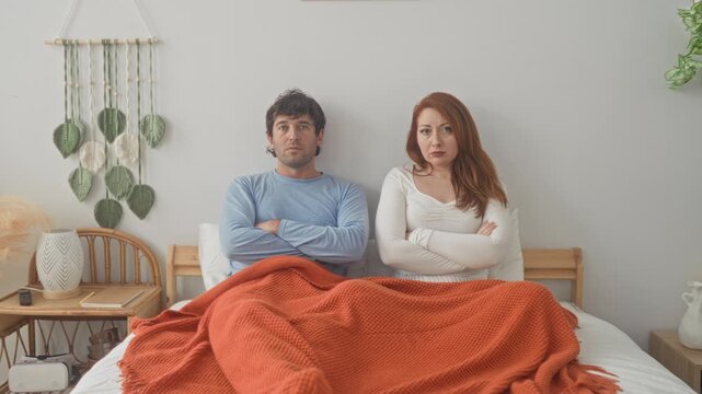 Man and woman sit upright on bed with arms crossed and orange blanket over legs in a cozy bedroom, both smiling toward camera; playful intimacy.