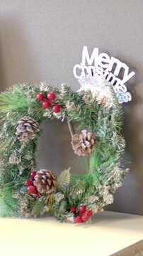 Vertical video: Panning camera revealing square wreath on tabletop and monitor, Merry sign blurring