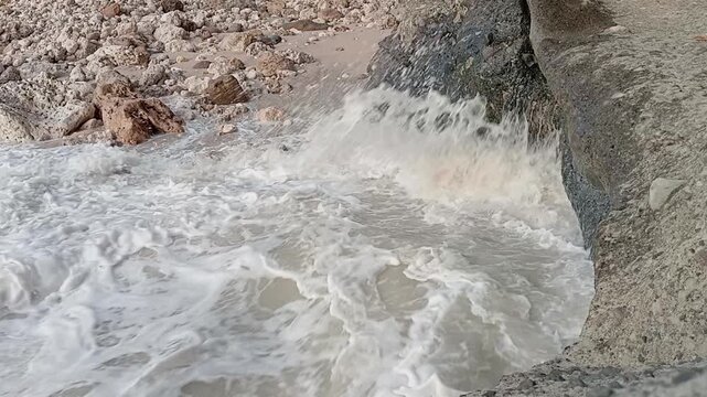 Slow motion or scenic video of heavy sea water and white seafoam crashing against big stones and rocky beach shoreline with natural coastal cliffs.
