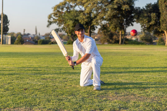 Male cricketer kneeling on grass holding bat, tracking red ball, wearing white uniform and dark cap