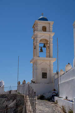 View of a traditional white bell tower with a blue dome and bronze bells against a clear blue sky with a person sitting on a stone path in Fira, Greece.