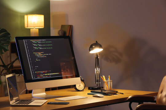 Programmer's workplace with computer, laptop and glowing lamp in dark office