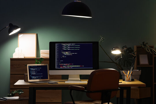 Programmer's workplace with laptop, computer and glowing lamps in dark office