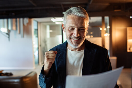 Smiling mature businessman celebrating success while reading document in modern office