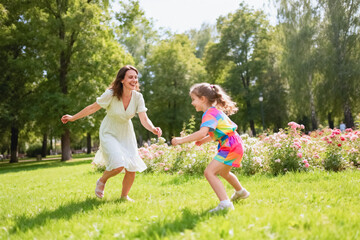 Fototapeta premium A joyful mother and her young daughter are playing and running together on a sunlit grassy park lawn surrounded by blooming roses and lush green trees.