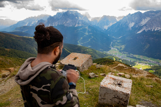 View of a man operating a drone remote controller on a mountain summit overlooking the jagged peaks of the Dolomites and a lush green valley Rocca Pietore, Veneto, Italy.