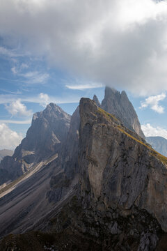 View of the jagged peaks of the Dolomites with steep rock faces and grassy ridges under a cloudy blue sky in Rocca Pietore, Veneto, Italy.