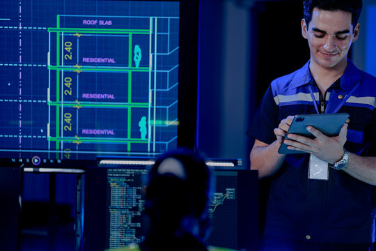 A construction engineer uses a tablet to review a BIM (Building Information Modeling) file. A CAD blueprint of a residential smart building is on a screen in the control center.