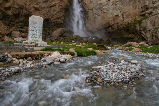 View of Khamosh Waterfall cascading down rugged cliffs into a rocky stream with a white monument in the foreground Skardu, Gilgit, Pakistan.