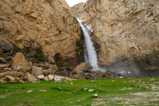 View of Khamosh Waterfall cascading down steep rocky cliffs into a green meadow with scattered boulders in Skardu, Gilgit, Pakistan.