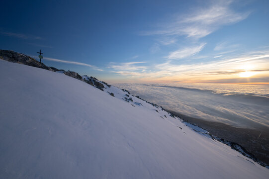 View of the snow-covered slope of Slavkovsky stit with a summit cross overlooking a sea of clouds at sunset in Vysoke Tatry, Presovsky kraj, Slovakia.