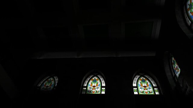Handheld video low angle interior view of a mosque featuring a series of arched stained glass windows with vibrant Islamic calligraphy and geometric patterns. The shot captures the dramatic contrast.