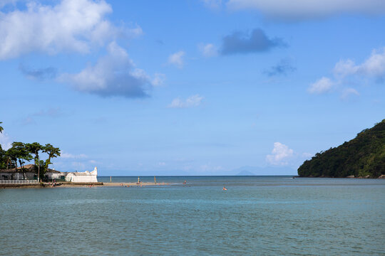 View of Bertioga channel entrance featuring historic fort, calm sea, palm trees, and coastal landscape under blue sky in Brazil.
