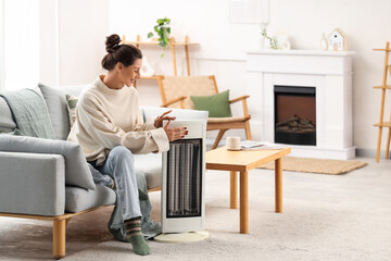 Fototapeta premium Young woman with electric heater sitting on sofa at home