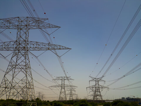 View of high-voltage power lines and transmission towers standing against a clear blue sky over desert vegetation UAE.