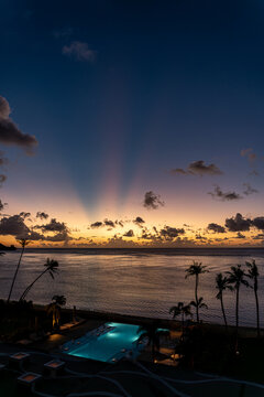 View of the sunset over Tumon Bay with palm trees and an illuminated swimming pool under a twilight sky with crepuscular rays in Tumon, Guam, Guam.