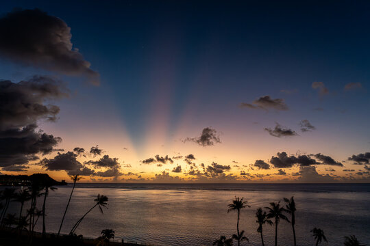 View of Tumon Bay at sunset with crepuscular rays stretching across the sky and palm trees silhouetted against the horizon in Tumon, Guam.