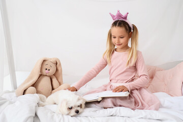Naklejka premium Cute little happy girl with Maltese dog and toy reading book on bed at home