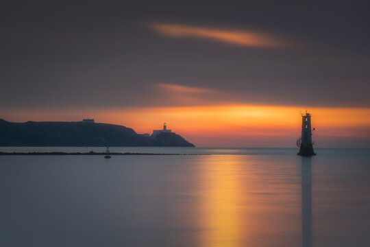 View of the South Wall lighthouse at sunset with calm water reflections and the silhouette of Howth Head in the distance, County Dublin, Ireland.