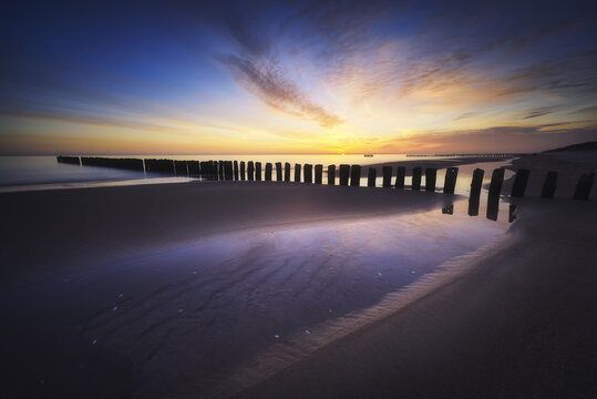 View of wooden breakwaters on a sandy beach under a colorful sunset sky with reflections on the water Chalupy, Wojewodztwo pomorskie, Poland.