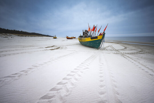 View of colorful wooden fishing boats with flags resting on the white sandy beach under a cloudy sky in Debki, Pomeranian Voivodeship, Poland.