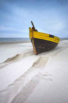 View of a yellow and brown wooden fishing boat resting on a sandy beach with deep tracks in the sand under a cloudy sky in Debki, Pomeranian Voivodeship, Poland.