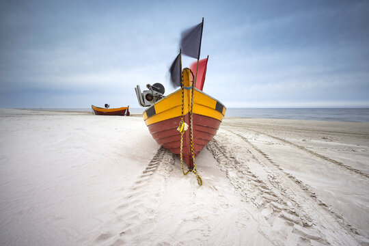View of a colorful traditional fishing boat with flags resting on the sandy beach under a cloudy sky in Debki, Pomeranian Voivodeship, Poland.