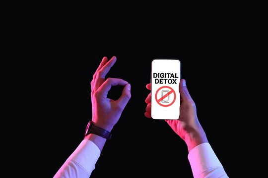 Hands holding smartphone with text DIGITAL DETOX on screen and showing OK against black background