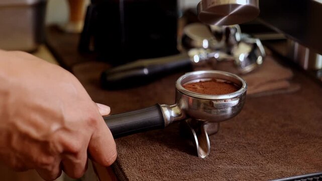Close-up of a barista's hands pressing ground coffee with a wooden-handle tamper into a stainless steel portafilter, preparing espresso shot for a professional coffee machine.