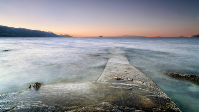 View of a stone pier extending into the calm Adriatic Sea with distant mountains under a soft sunset sky in Croatia.