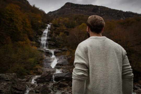 View of a man in a knitted sweater looking at a cascading waterfall surrounded by autumnal trees and rocky cliffs in Verzasca, Ticino, Switzerland.