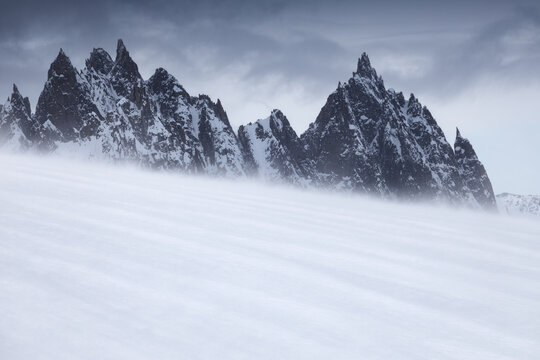 View of jagged dark mountain peaks rising above a vast wind-swept snow field under an overcast sky in Aosta Valley, Italy.