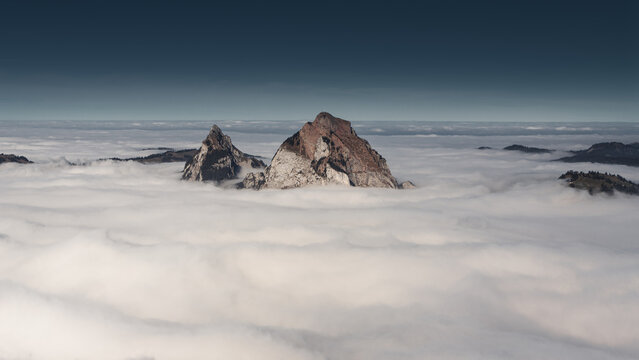 View of the Mythen mountain peaks rising above a thick sea of clouds under a dark blue sky near Stoos Morschach, Schwyz, Switzerland.