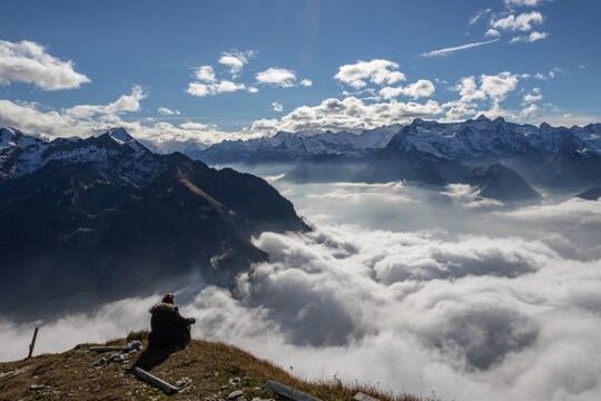 View of a person sitting on a grassy ridge at Stoos overlooking a thick sea of clouds and snow-covered mountain peaks under a bright blue sky in Morschach, Schwyz, Switzerland.