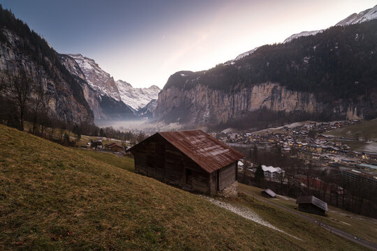 View of a traditional wooden barn on a grassy hillside overlooking the valley village and steep cliffs at dusk in Lauterbrunnen, Canton of Bern, Switzerland.