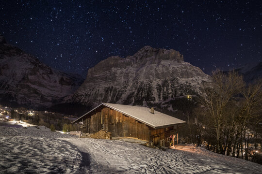 View of a traditional wooden chalet in a snowy landscape under a starry night sky with snow-capped mountains in Grindelwald, Canton of Bern, Switzerland.