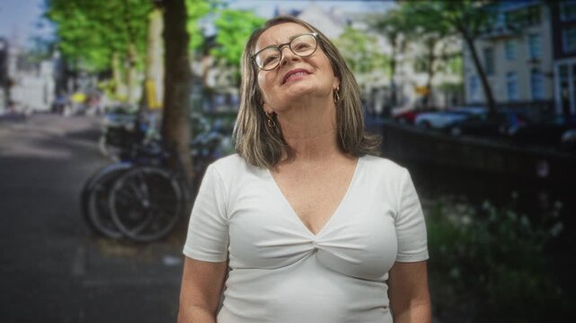 Woman with grey hair and glasses tilts head back showing cleavage in a white top on a city street beside bicycles and a canal; contentment reflection calm.