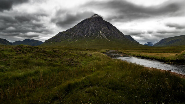 View of the iconic Buachaille Etive Mor mountain peak rising above a winding river and green moorland under a dramatic cloudy sky in Glencoe, Scotland, United Kingdom.