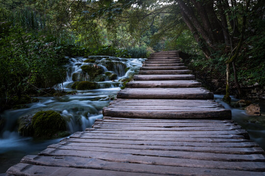 View of a wooden boardwalk path winding through lush forest beside a cascading waterfall over mossy rocks in Plitvice Lakes National Park, Plitvica Selo, Lika-Senj County, Croatia.