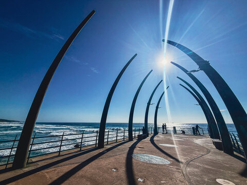 View of the Whalebone Pier featuring large curved structures and people walking under a bright sun at uMhlanga, KwaZulu-Natal, South Africa.