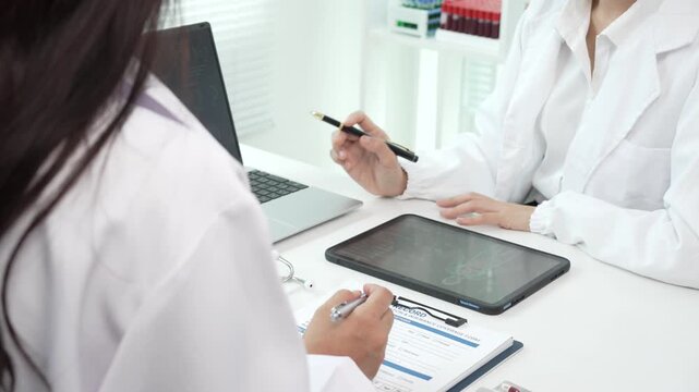 Two doctors in white coats review medical notes and a digital tablet at a clinic desk, showing teamwork, consultation, and modern healthcare in a bright office.